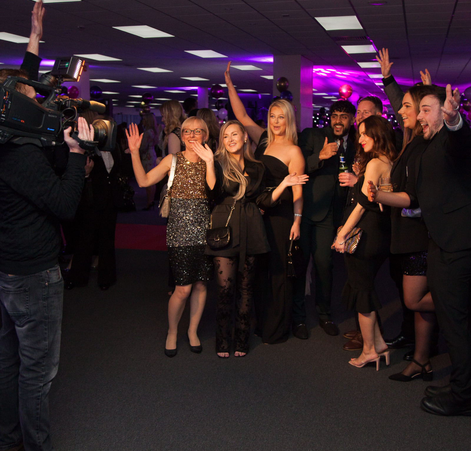 A group of eight people poses for a photo at an event, with some raising their hands in excitement. They are dressed in formal and semi-formal attire, and a cameraman is capturing the moment in the foreground. The backdrop features purple lighting and decorations, creating a festive atmosphere.