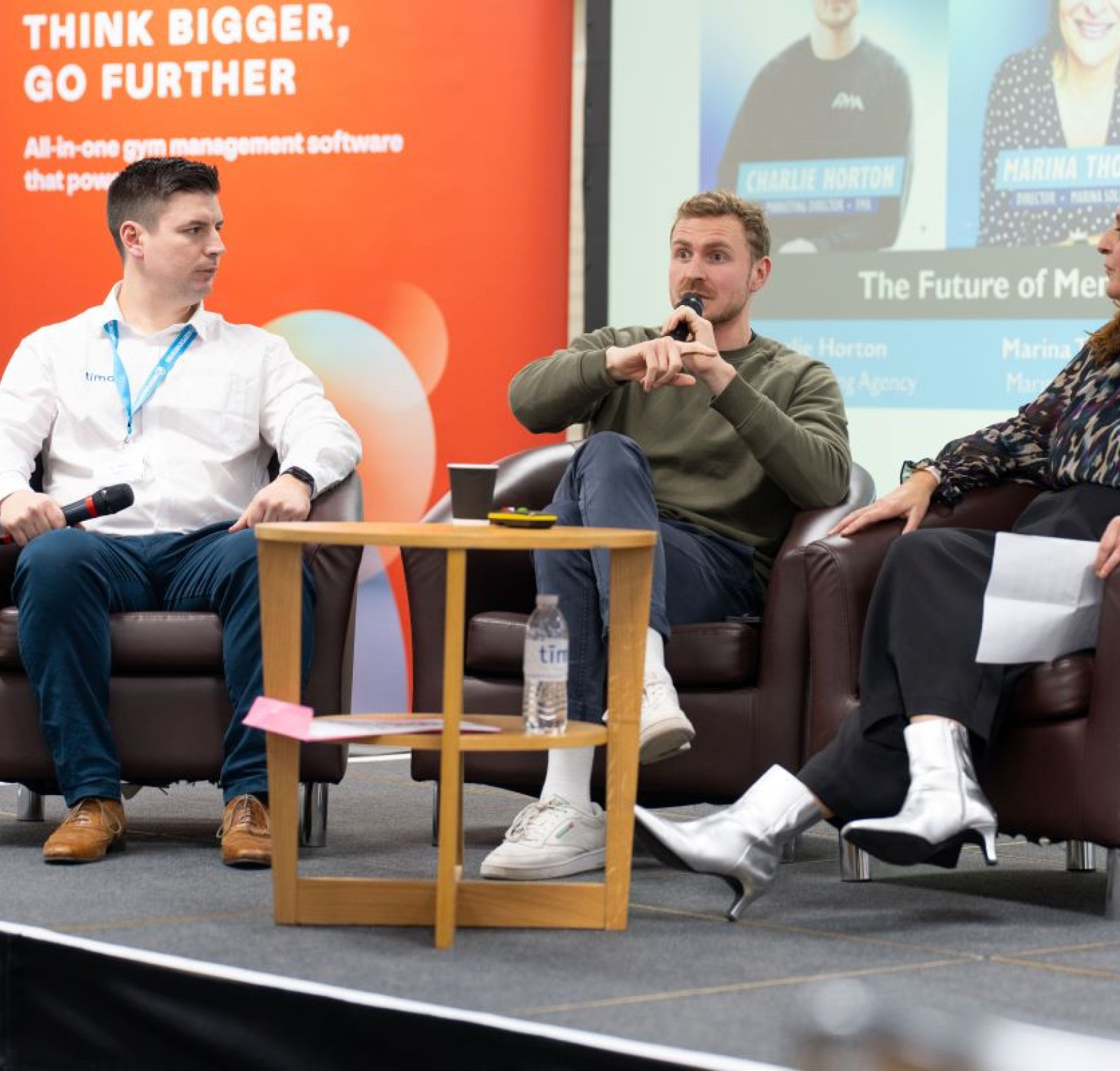 A panel discussion featuring three speakers sitting in armchairs on a stage. The background displays promotional banners and a presentation screen. The central speaker is gesturing while talking, with two other individuals, one on each side, listening attentively. The audience is not visible.