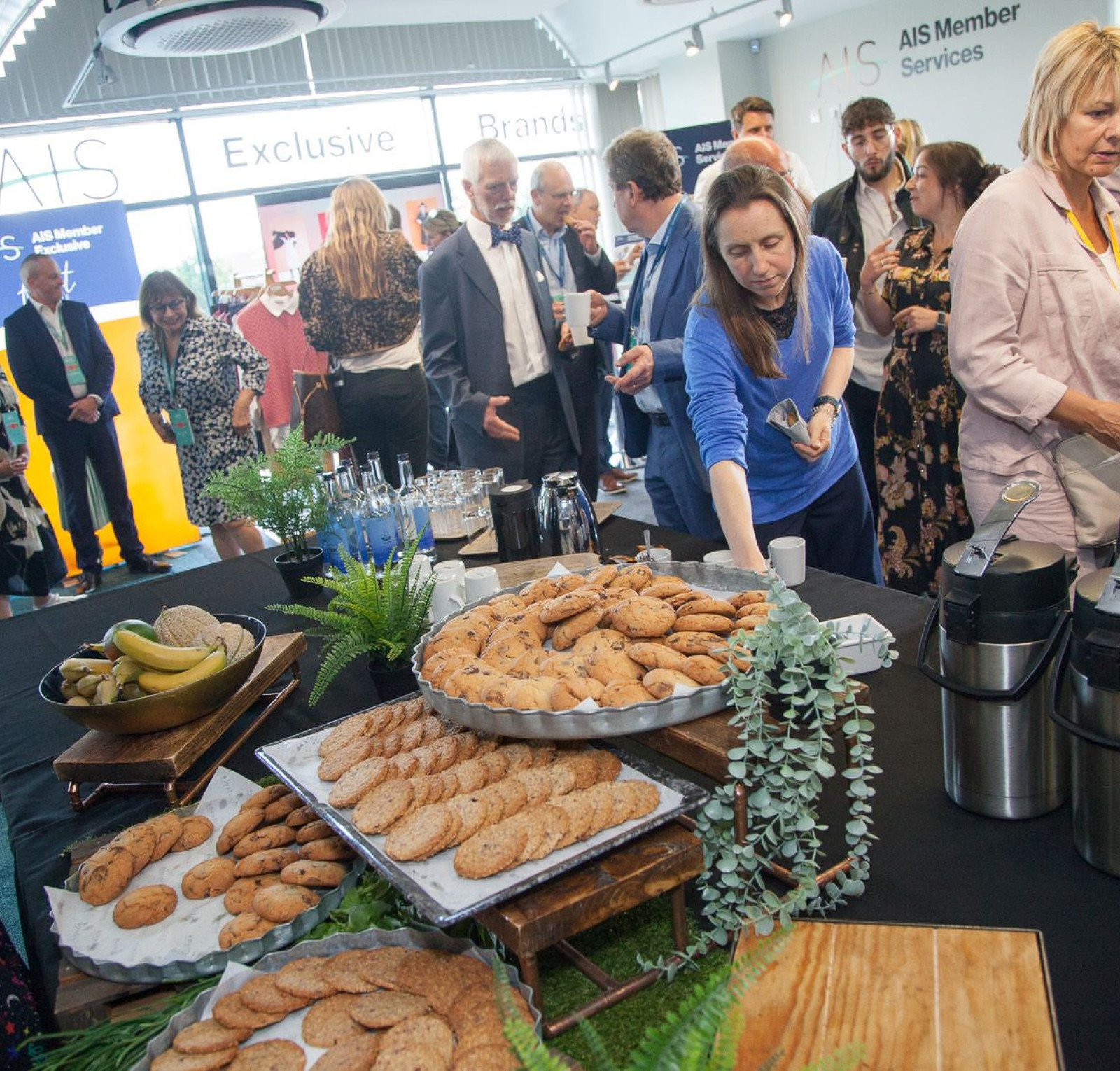 A busy conference setting with a diverse group of attendees networking around a table laden with various cookies and snacks. Some people are engaging in conversation, while one woman in a blue shirt serves food. The background features branding banners and a modern event space.