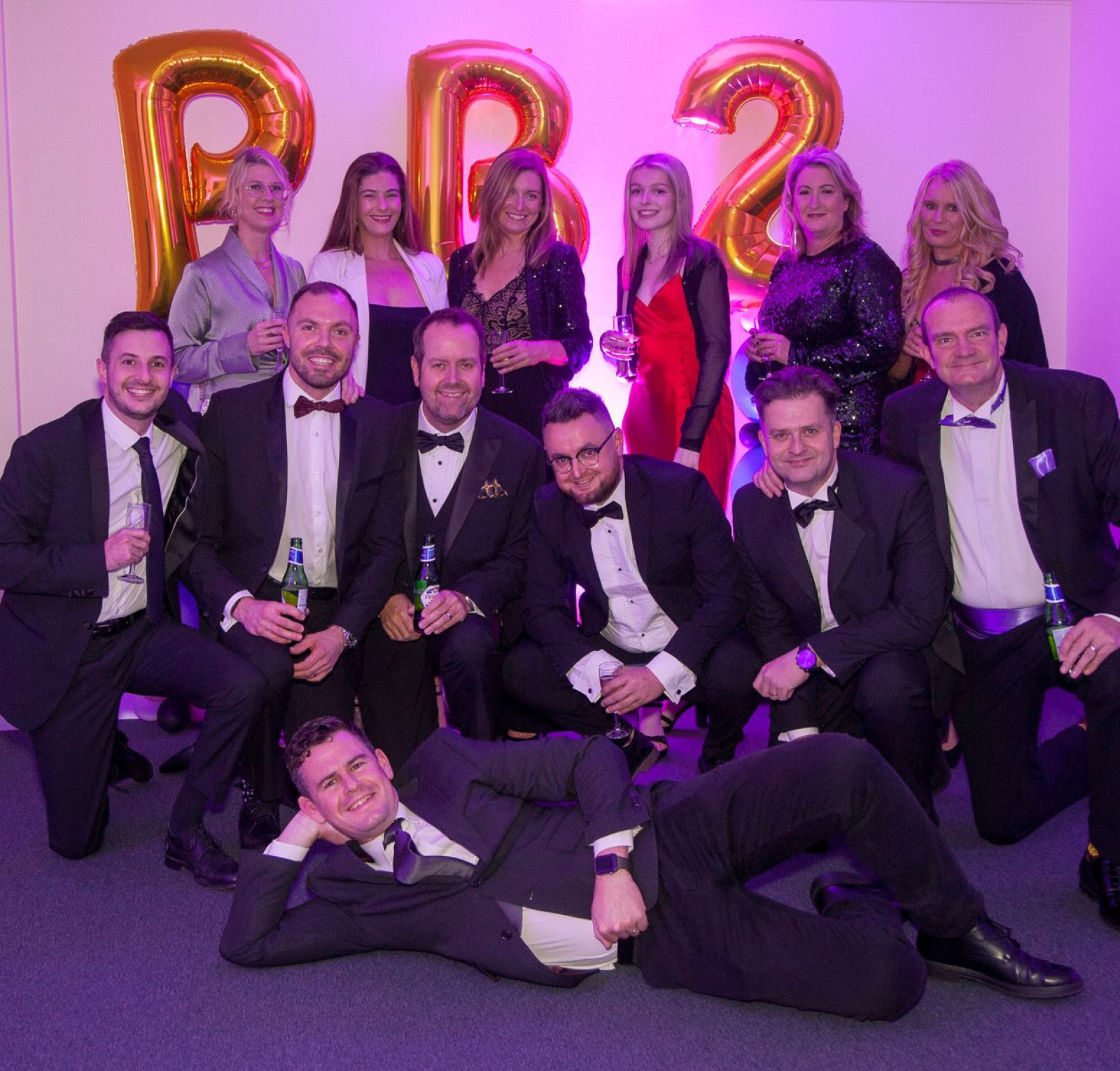A group of men and women dressed in formal attire poses for a photo at a celebration. They are holding drinks and smiling, with large decorative 
