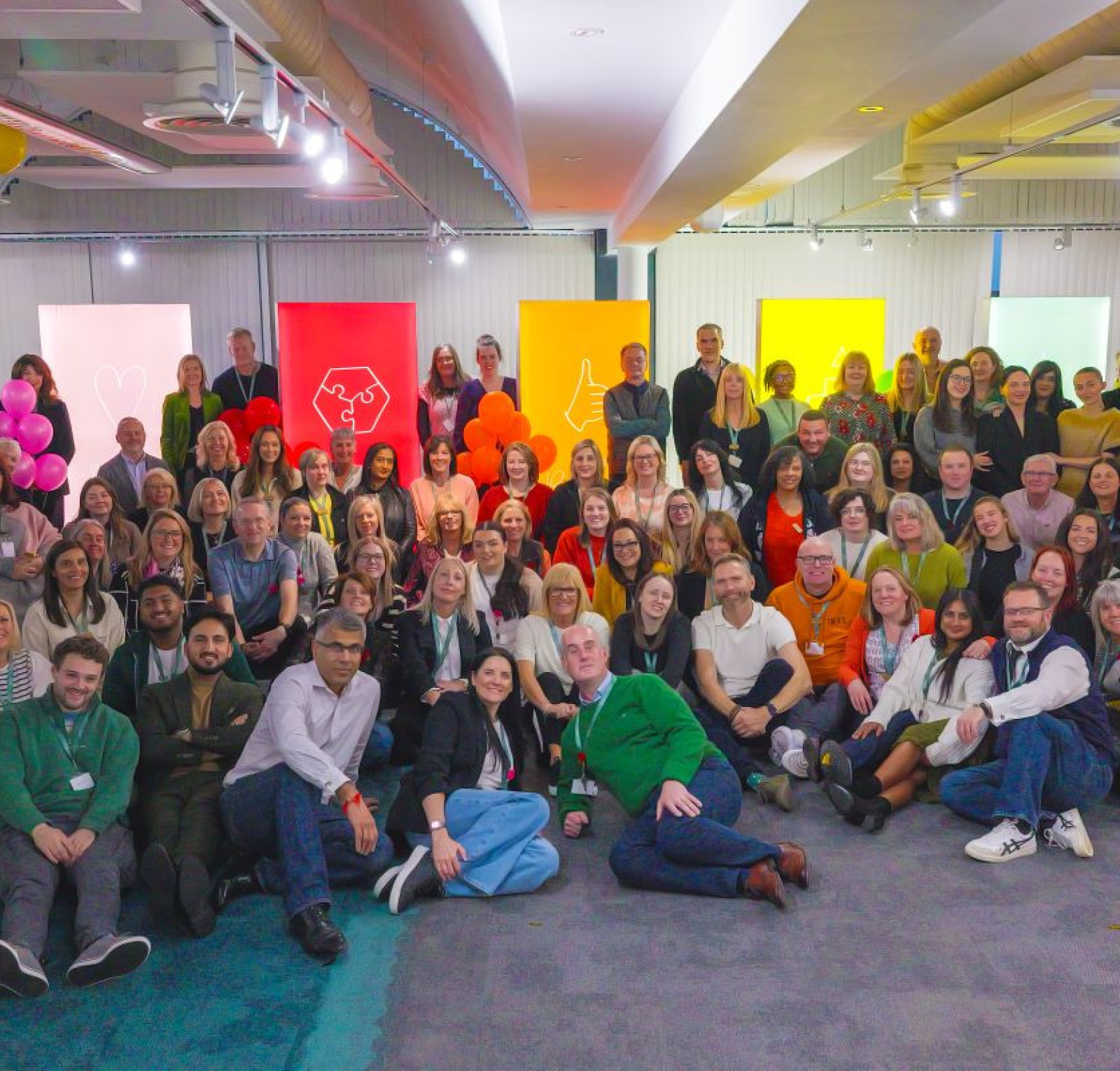 A large group of diverse people gathers for a photo in a brightly lit room decorated with balloons and colorful posters. They sit and stand in a relaxed pose, smiling at the camera.