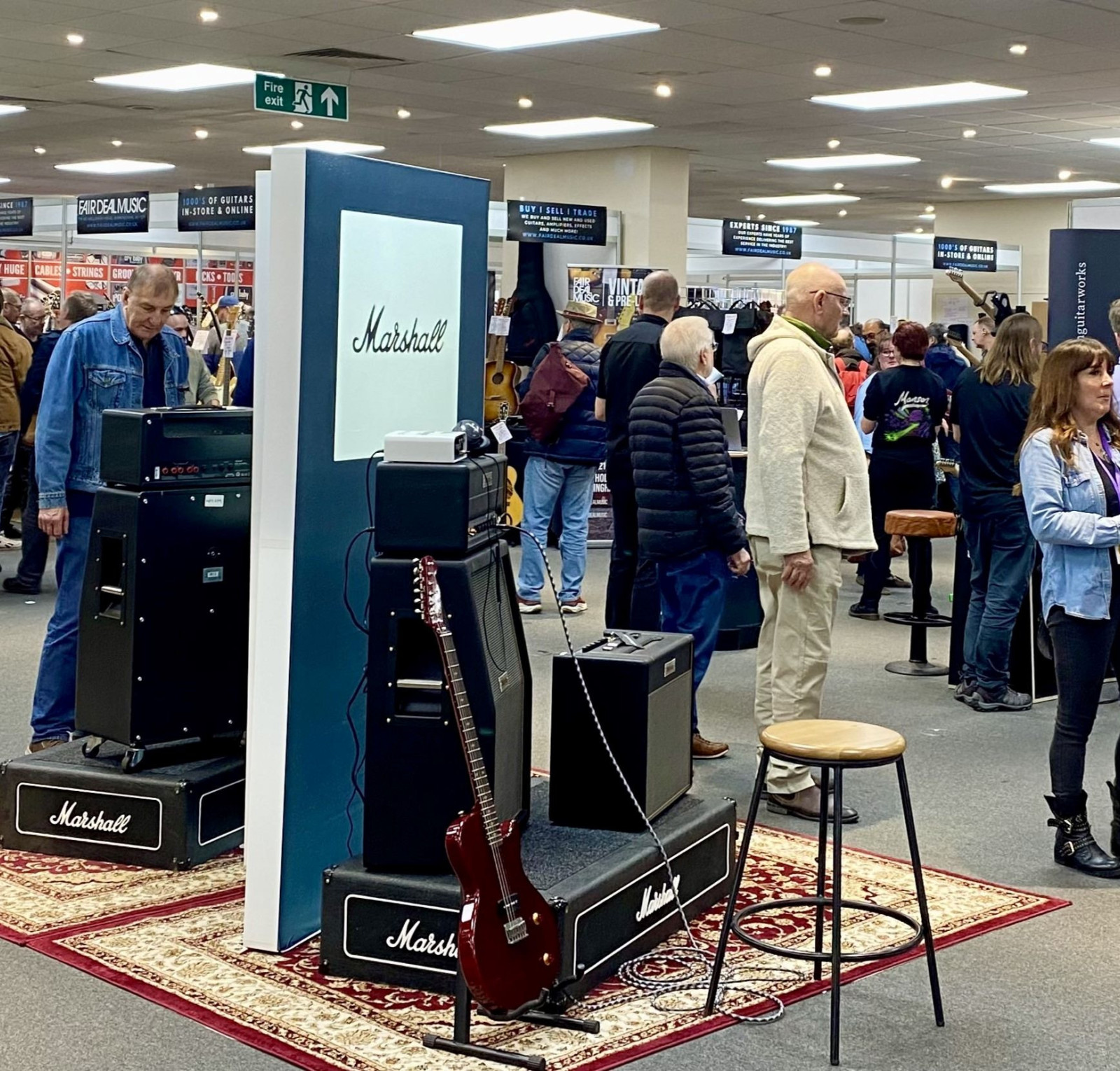 A bustling exhibition space filled with people exploring various stands. In the foreground, there are Marshall guitar amplifiers and a guitar on display, surrounded by attendees engaging with the booths and merchandise.