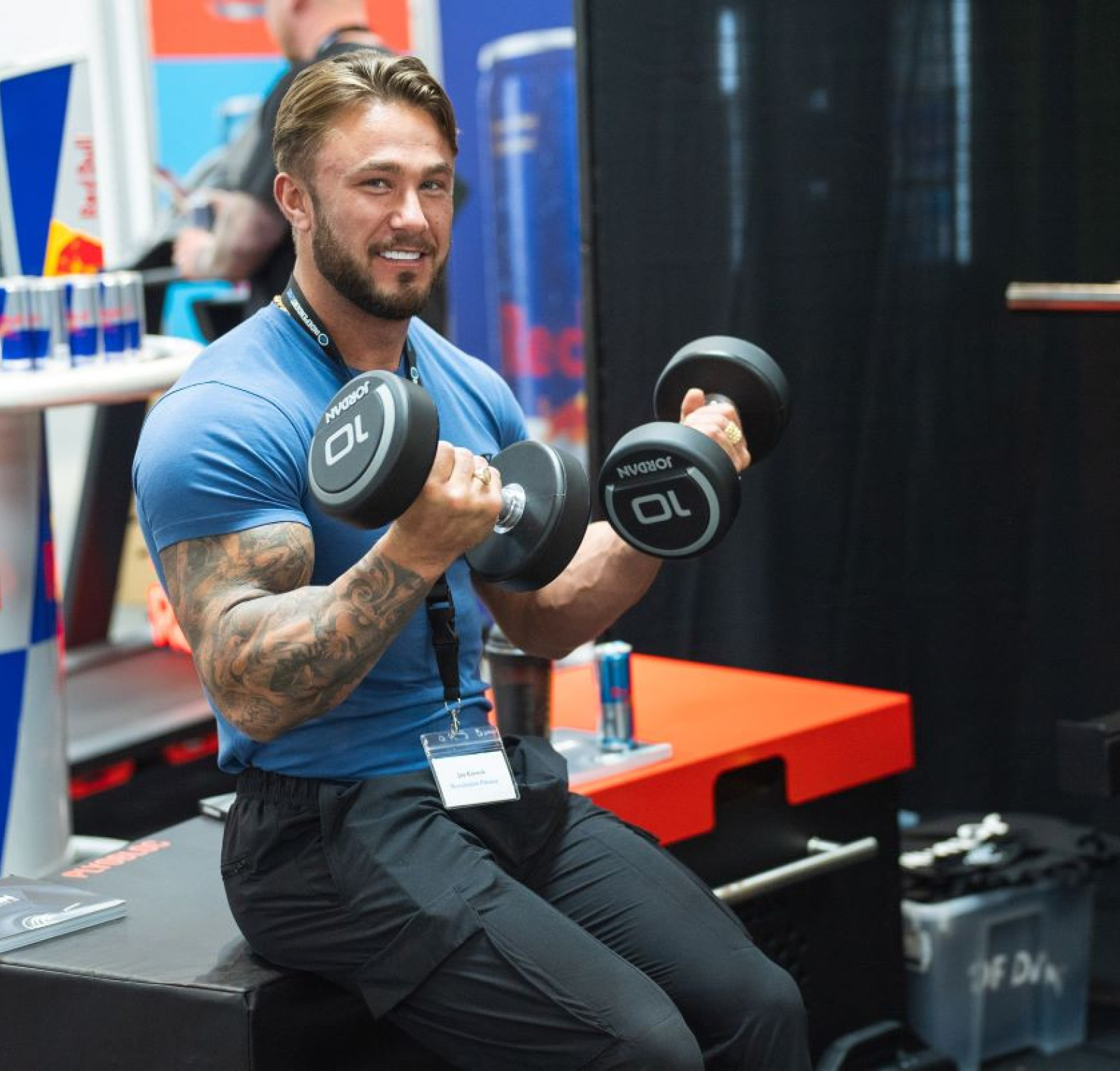 A muscular man with tattoos smiles while lifting dumbbells on a bench. He wears a blue shirt and black pants. In the background, there are Red Bull energy drink cans and promotional materials.