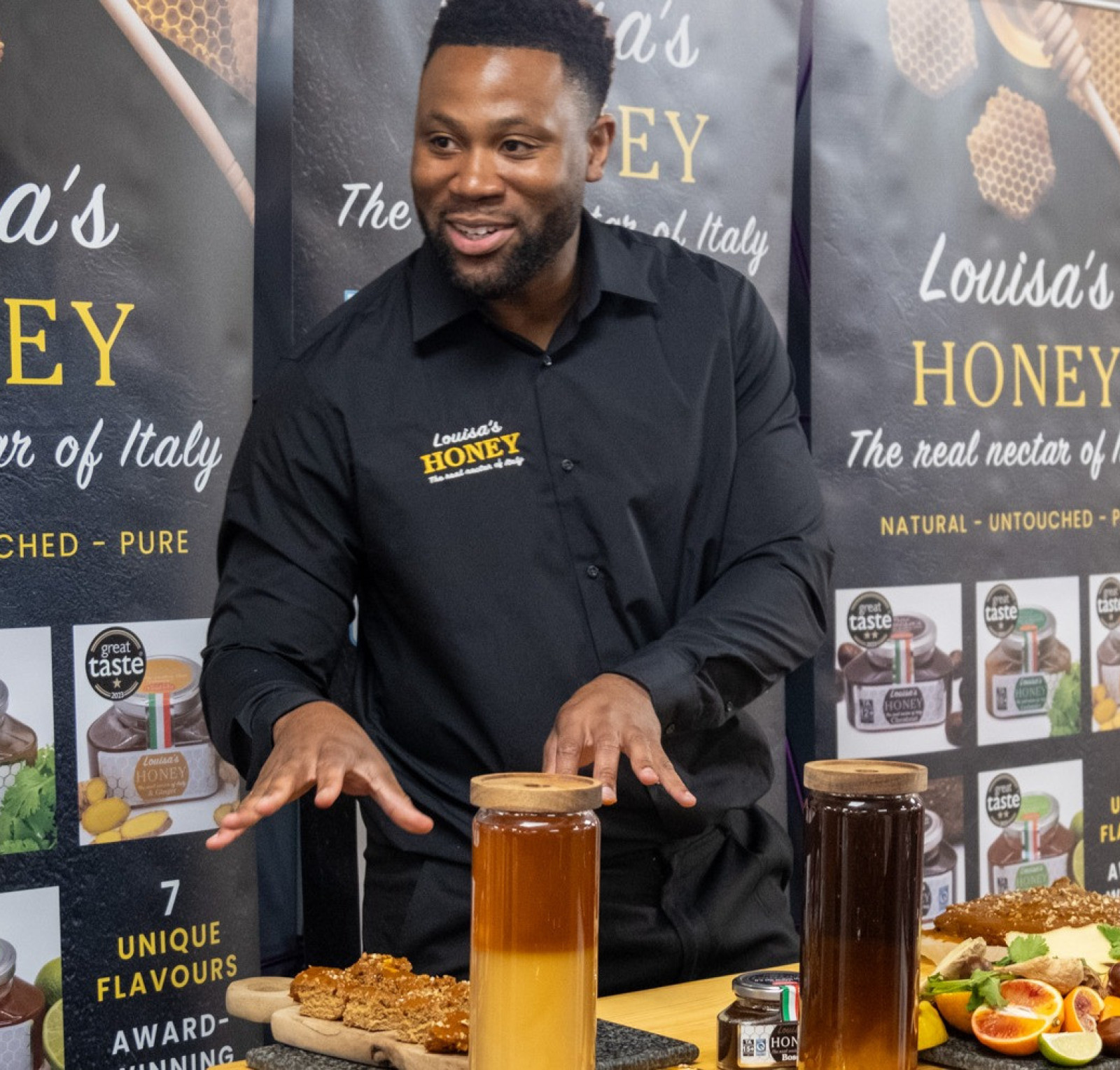 A man in a black shirt gesturing enthusiastically stands behind a table with various honey jars and honeycomb. The background features banners promoting Louisa's Honey, highlighting its purity and unique flavors. Colorful fruits are displayed on the table.