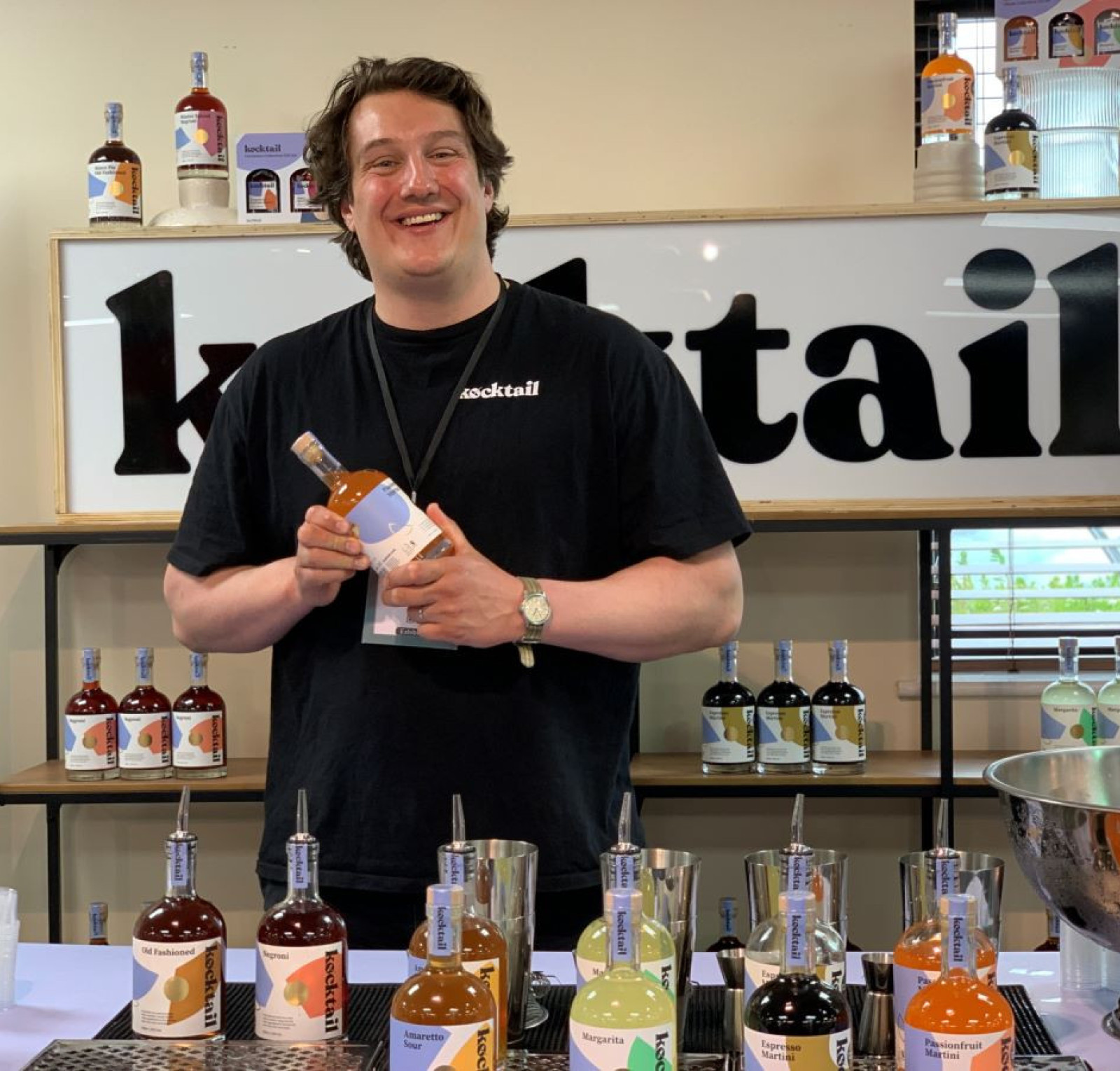 A smiling bartender holds a bottle of cocktail mix, standing behind a table lined with various bottles of colorful beverages. A large sign reading 
