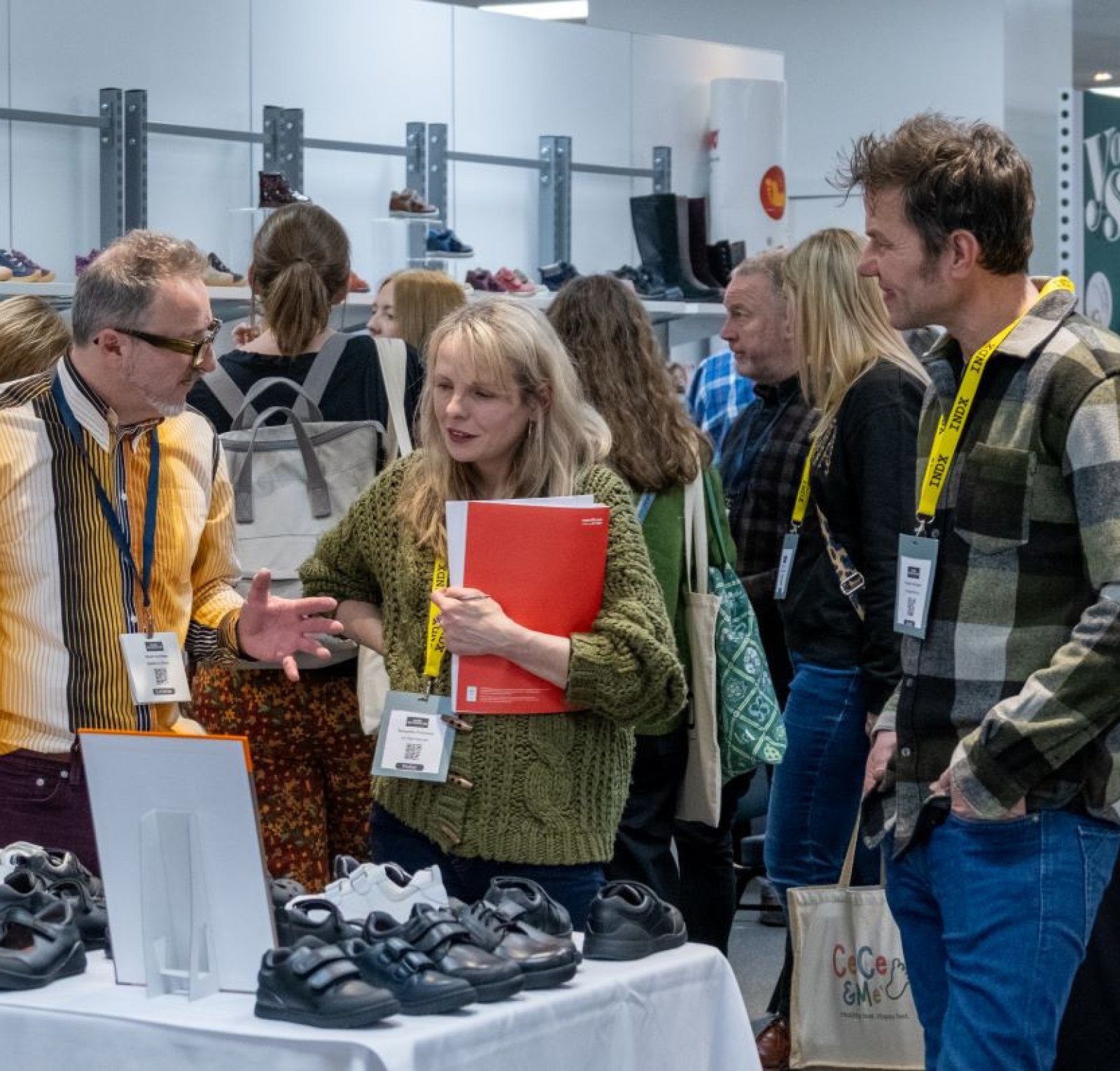 A group of people is discussing footwear at a trade show. One man with glasses passionately explains details about shoes, while a woman holding a red folder listens intently. Another man looks on with interest. The display features various styles of shoes in the background.