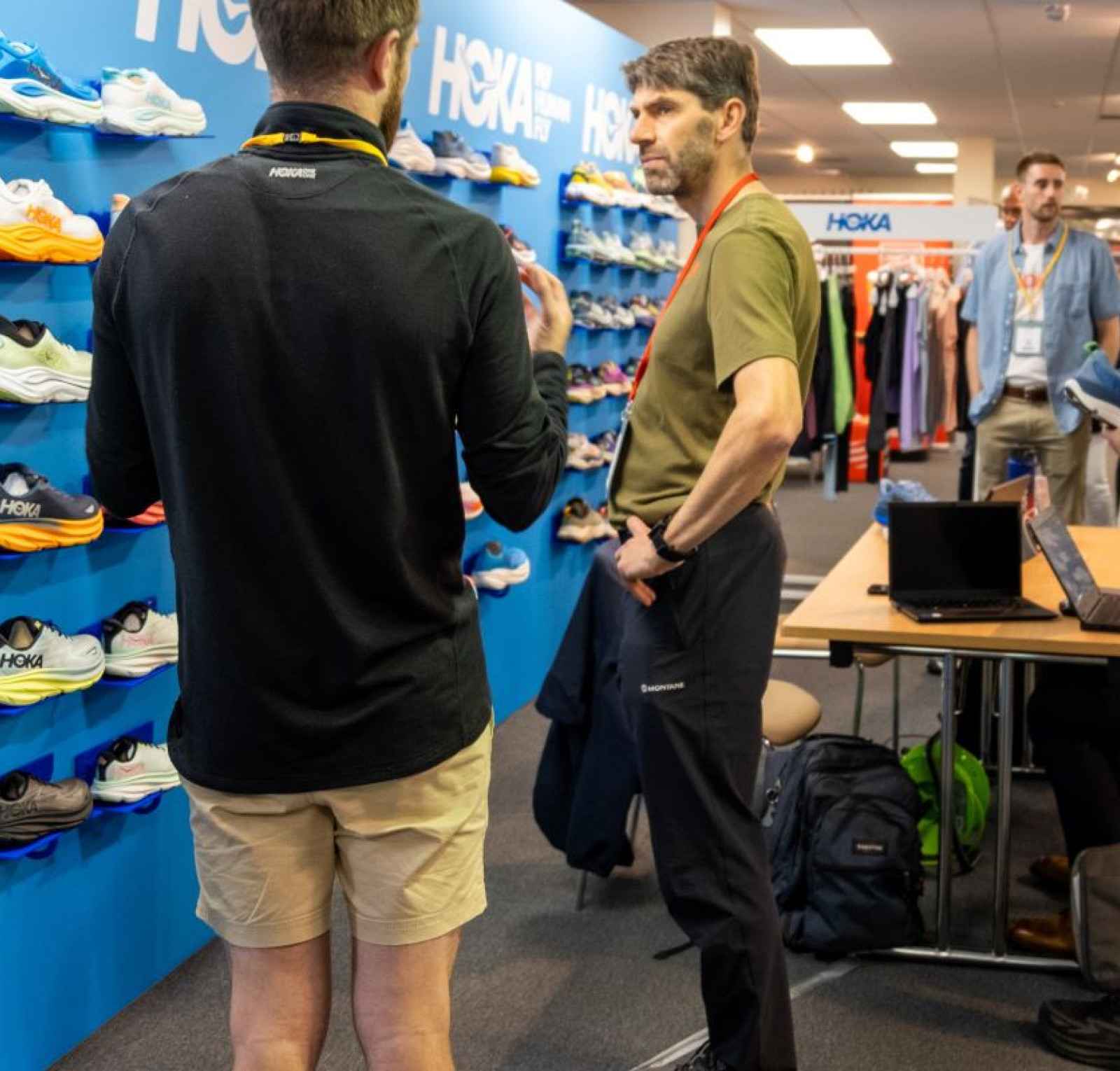 A group of people engage in conversation at a shoe display featuring various HOKA athletic shoes. Two individuals in the foreground are talking, while others browse the display or work at a table with a laptop. The atmosphere is casual, with colorful shoes prominently showcased in the background.