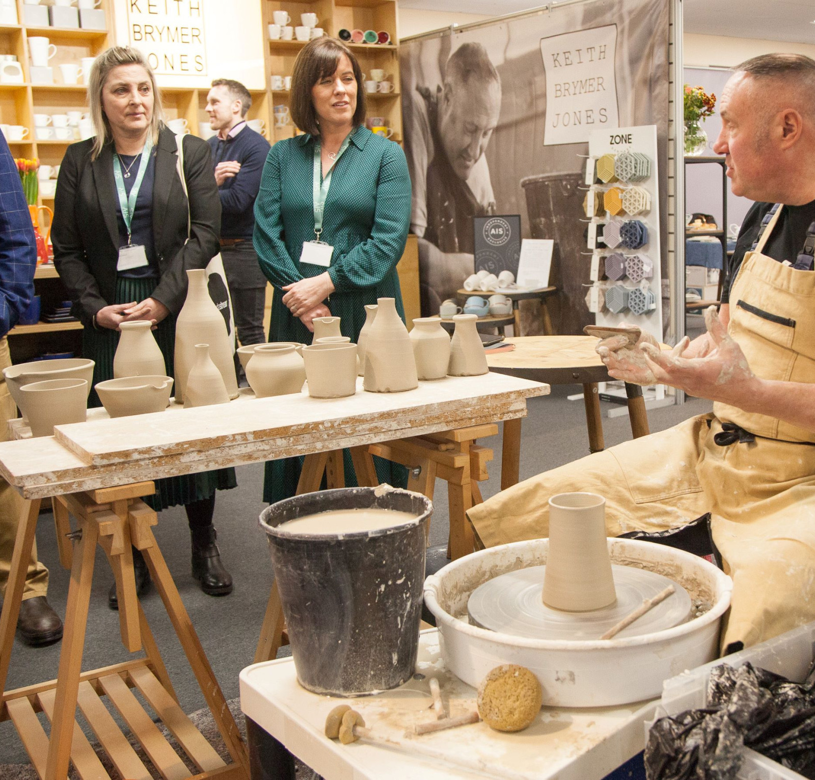 A potter demonstrates their craft at a workshop, shaping clay on a wheel. Several attendees watch attentively, including two women and two men. A table with various clay pots and tools is prominently displayed in the foreground.