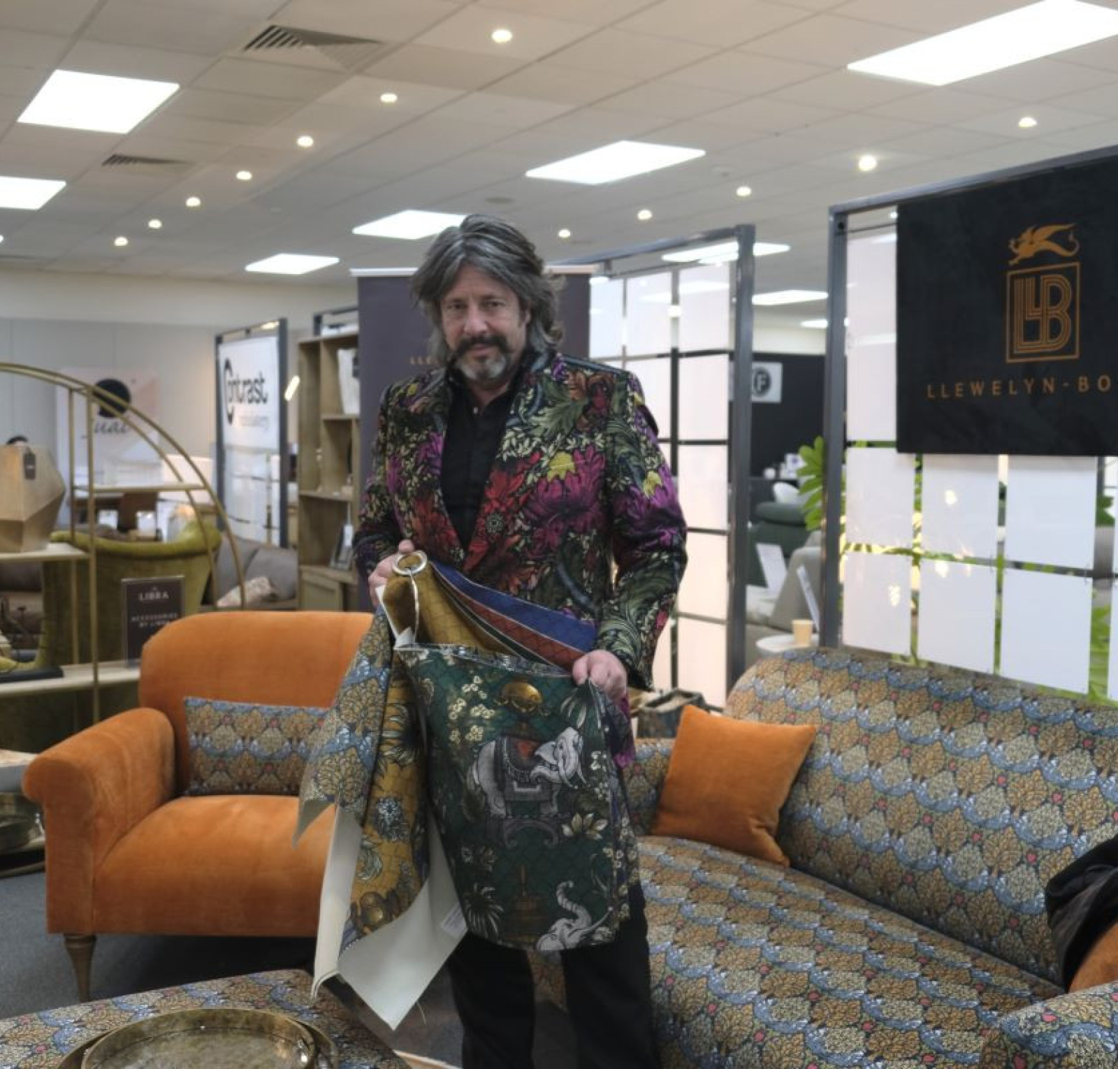A man stands in a stylish interior filled with furniture and decorative items, wearing a colorful, patterned jacket. He holds an elegant fabric with intricate designs, showcasing a blend of modern and classic decor elements.