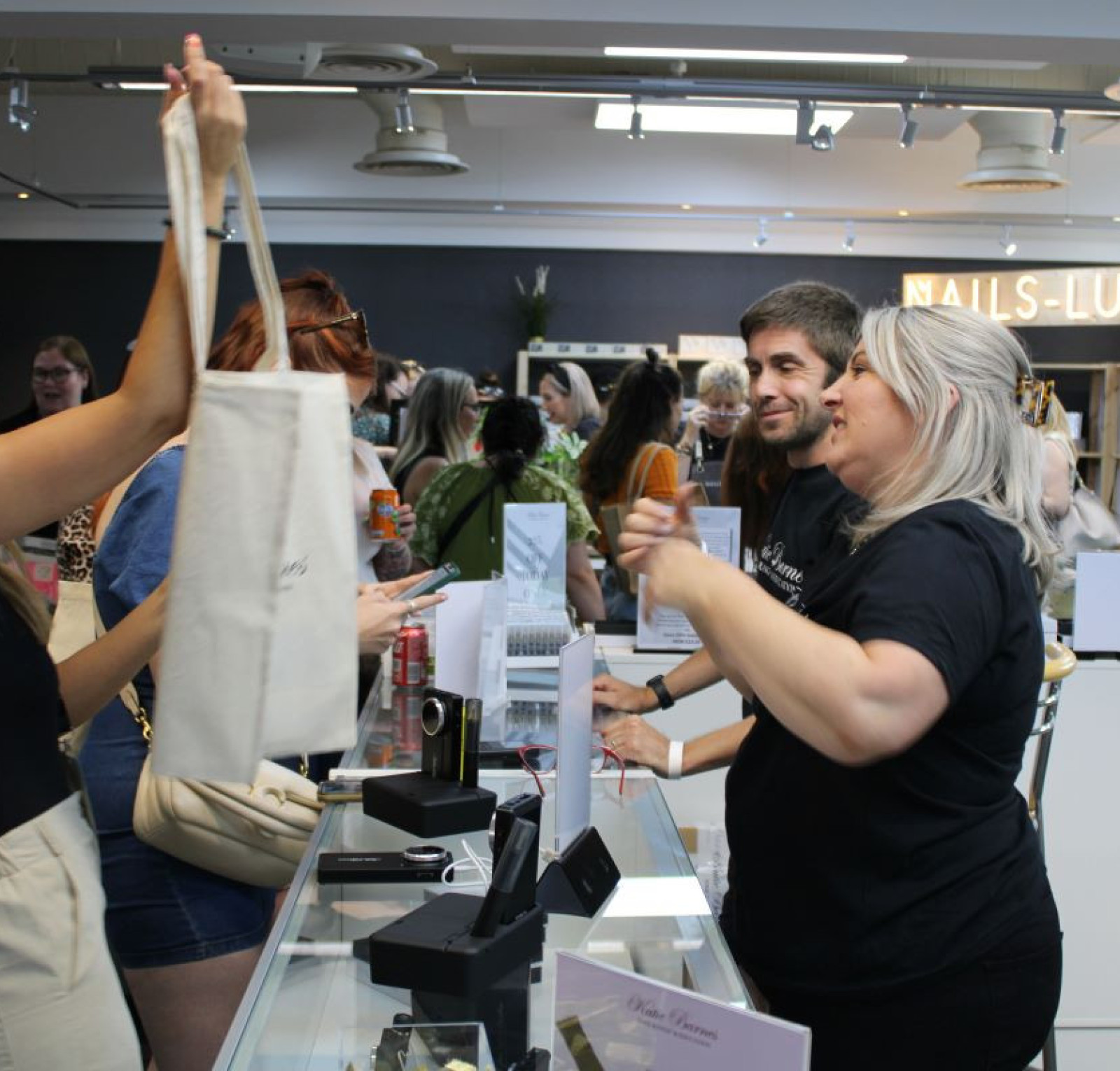 A lively scene at a cosmetics store with customers interacting with staff. A woman happily holds a bag while another staff member assists her. Several shoppers browse the displays in the background, creating a bustling atmosphere.