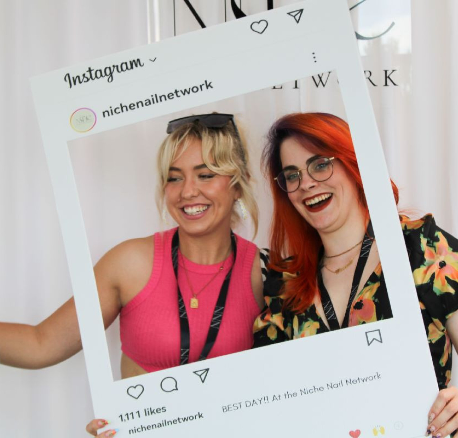 Two young women pose together, smiling and holding a large Instagram-style photo frame. One has blonde hair and is wearing a pink top, while the other has bright red hair and glasses. They are at an event, and banners in the background indicate the niche nail network.