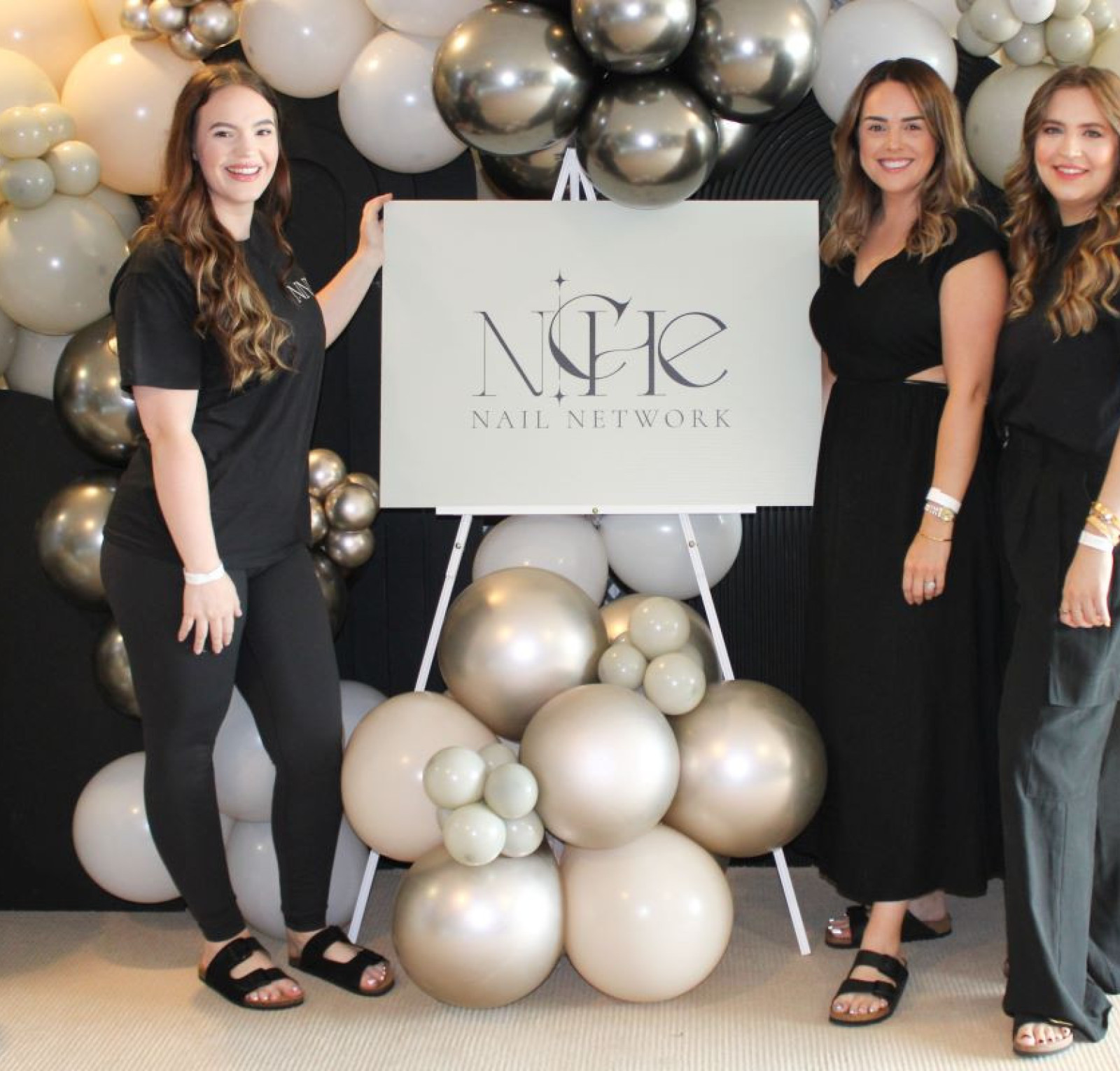 Three women stand in front of a decorative balloon arch, smiling beside a sign for 