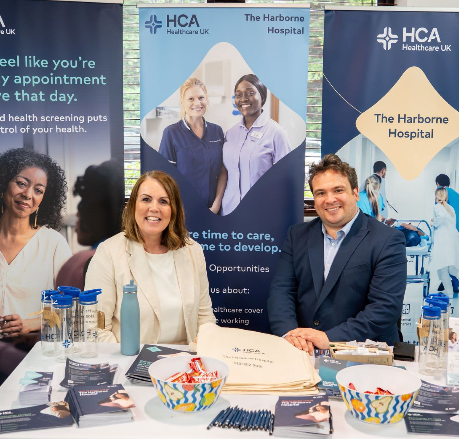 A healthcare event booth featuring two professionals smiling at the camera, surrounded by informational banners about HCA Healthcare UK and The Harborne Hospital. Promotional materials, a water bottle, and a tote bag are visible on the table.