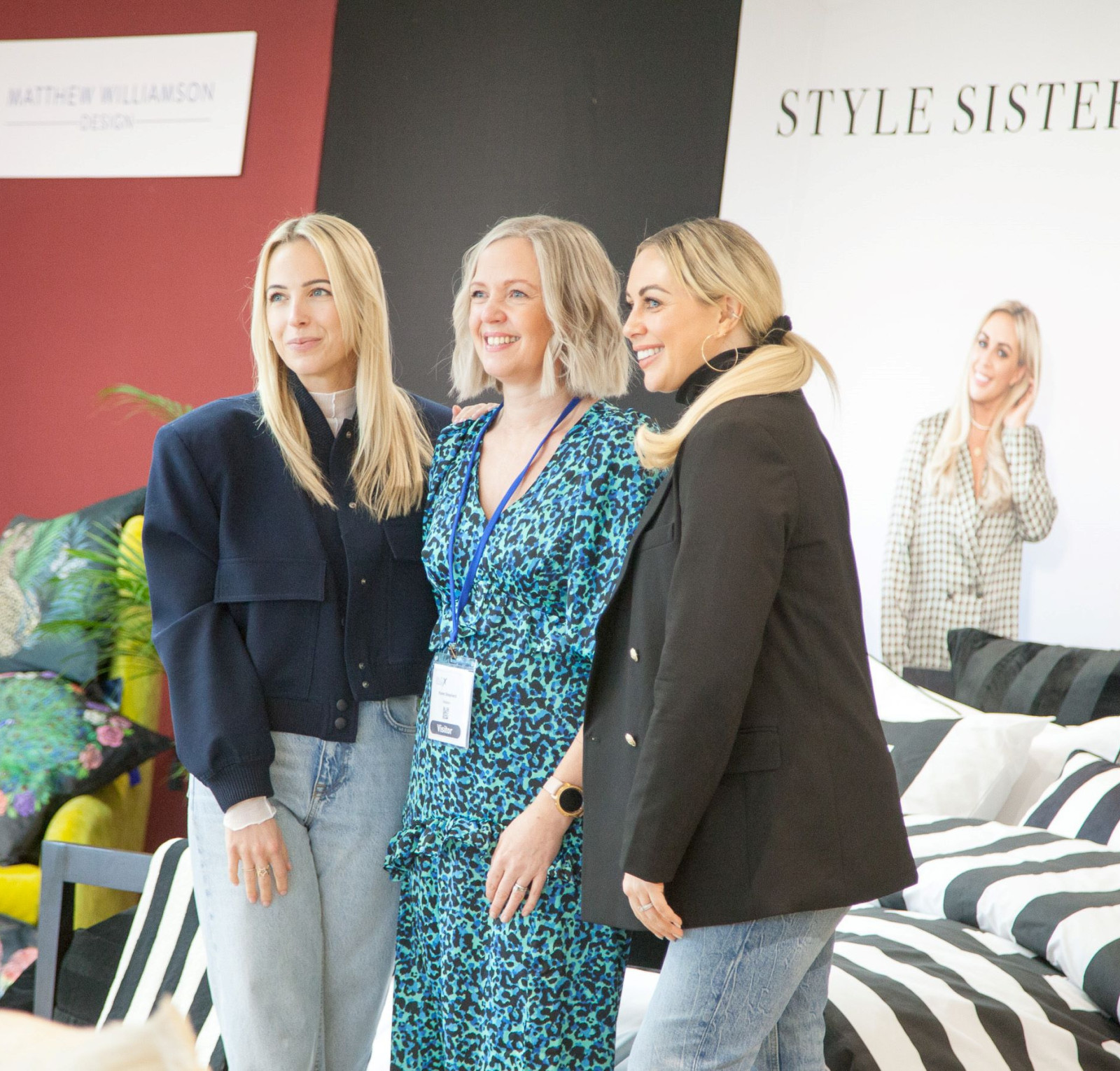 Three women pose together at an event, smiling. One woman wears a black jacket and blue jeans, another is in a blue floral dress, and the third has blonde hair in a ponytail with a black blazer. Colorful furniture and patterned fabrics are displayed in the background.
