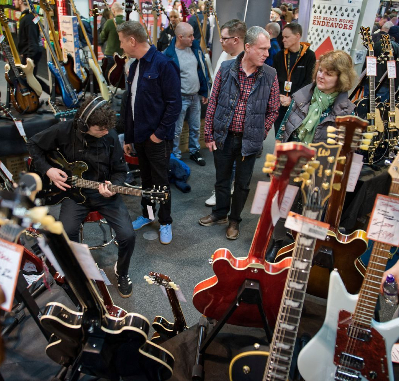 A busy music trade show with people interacting around numerous guitars displayed on tables. A guitarist is seated, playing an electric guitar while onlookers watch. Various guitar models and brands are visible in the foreground and background.