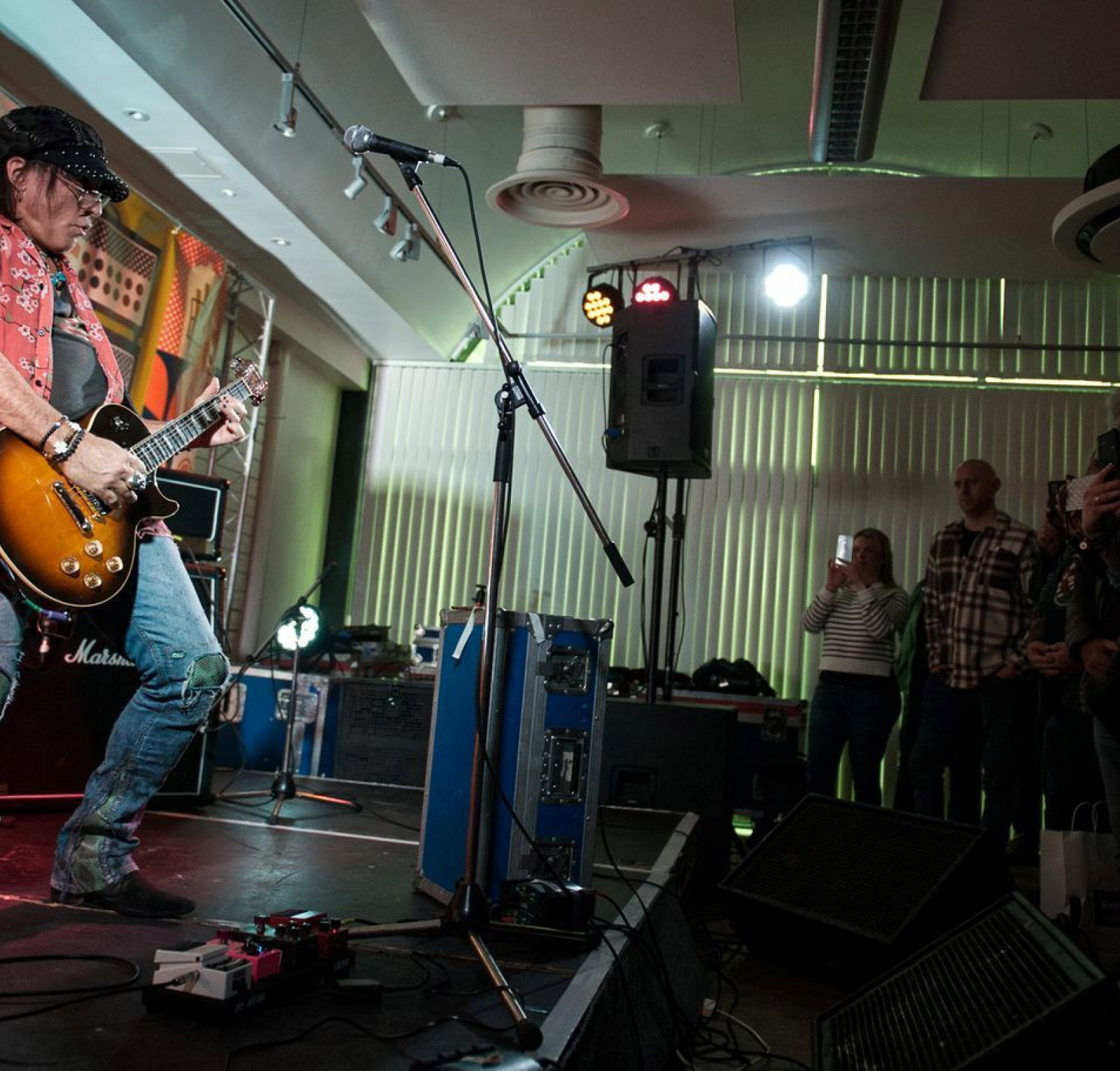 A guitarist in a black cap and a sleeveless shirt performs passionately on stage, playing an electric guitar. Colored lights illuminate the scene, and an engaged audience watches from the foreground. Amplifiers are visible behind the performer.