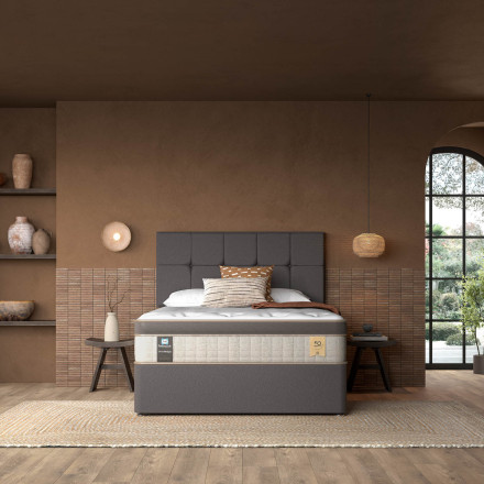 A stylish bedroom featuring a modern bed with a gray upholstered headboard. The bed is dressed in neutral bedding, complemented by decorative pillows. Surrounding the room are earthy tones, with shelves displaying vases and art pieces. A large window lets in natural light, revealing greenery outside. A woven rug adds texture to the wooden floor.