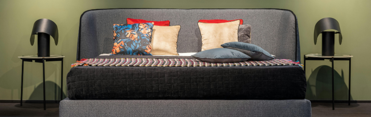 A modern bed with a gray upholstered headboard featuring colorful pillows in red, gold, blue, and floral patterns. Two side tables with sleek black lamps flank the bed against a green wall. A patterned throw is placed at the foot of the bed.