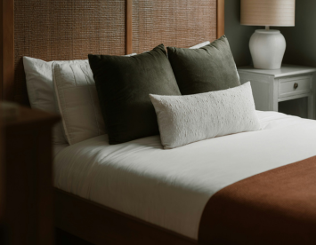 A cozy bedroom featuring a neatly made bed with a white duvet, adorned with several decorative pillows in shades of green and a white textured pillow. A warm brown throw blanket is placed at the foot of the bed, and a bedside table with a lamp is visible in the background.
