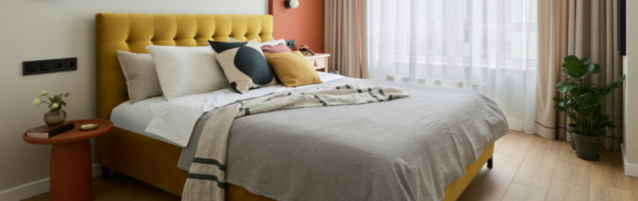 A modern bedroom featuring a yellow tufted headboard, a cozy bed with white and patterned pillows, and a gray blanket. There’s a small nightstand with decorative items and a window dressed with light curtains, allowing natural light to fill the room. A potted plant adds a touch of greenery.