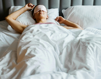 A person lying in bed, wearing a sleep mask and resting peacefully under white bedding.