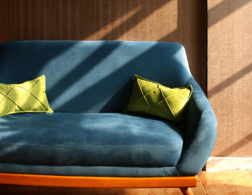 A modern blue sofa with two green decorative pillows is illuminated by natural light, casting shadows on a textured brown wall.