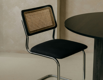 A modern chair featuring a black fabric seat and a backrest made from woven cane, positioned next to a round dark table against a textured beige wall.