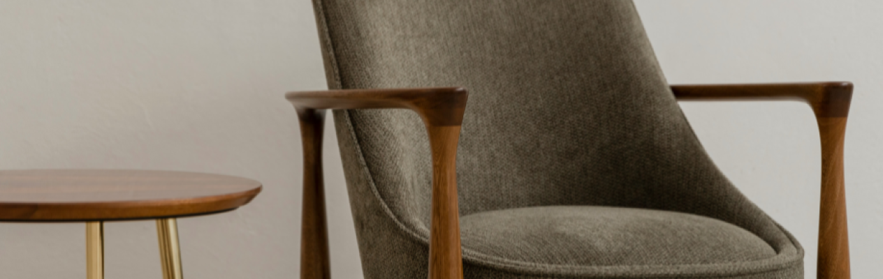 A modern, upholstered armchair in a muted green tone with wooden armrests sits next to a round wooden side table featuring gold legs. The background is a simple, neutral color.