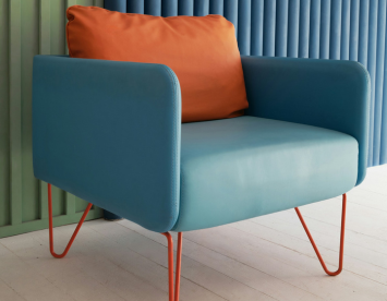 A modern armchair featuring a blue body and an orange cushion, resting on slender orange legs, set against a backdrop of vertical blue and green panels.