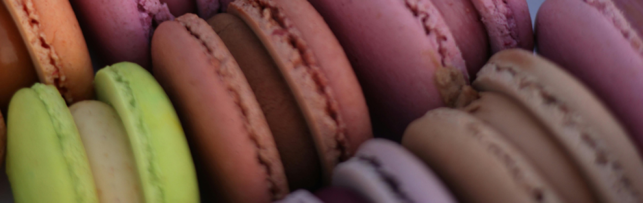 A variety of colorful macarons displayed in a circular arrangement, showcasing shades of yellow, orange, pink, purple, brown, and beige.