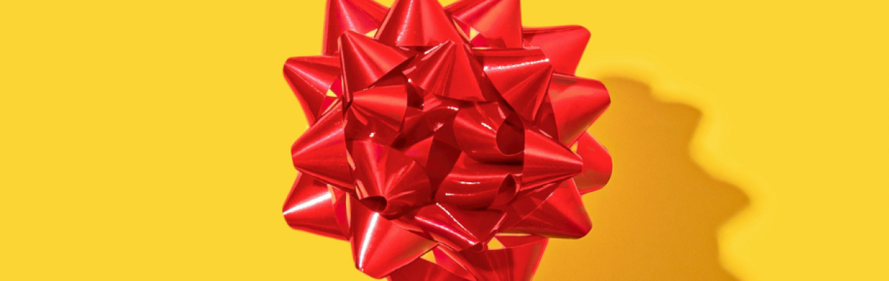 A shiny red gift bow with pointed ends, placed on a bright yellow background, casting a shadow.