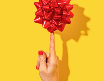 A hand with red nail polish is pointing at a shiny red gift bow against a bright yellow background. The bow's shadow is visible on the wall.