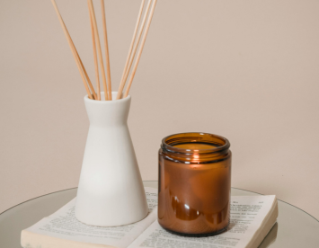 A white ceramic vase with bamboo reed sticks next to a small amber-colored glass candle jar, placed on an open book on a reflective surface.