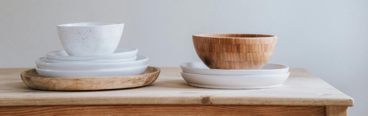 A stacked arrangement of white ceramic plates and bowls beside a wooden bowl on a light wooden table. The scene features a mix of textures with the smooth ceramic and the natural finish of the wood.