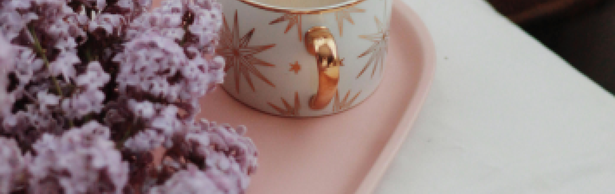 A delicate porcelain cup with gold accents sits on a pink tray alongside a bouquet of lilac flowers. The cup is filled with a warm beverage, likely coffee. Soft, neutral bedding provides a cozy backdrop.