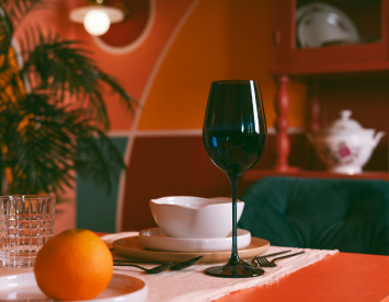A stylish dining table set with a blue wine glass, a white bowl, and plates, accompanied by a clear glass, silver utensils, and an orange. The background features colorful walls and decorative elements like a plant and a teapot.
