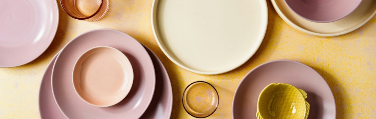 A collection of various ceramic plates and bowls in soft pastel colors, including pink, yellow, and green, arranged on a textured yellow background. A round, pale yellow plate is at the center, surrounded by multiple round and oval plates in shades of pink and purple, along with a small green dish and a glass.