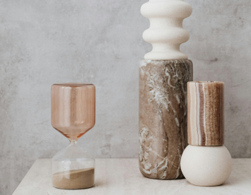 Three decorative vases arranged on a light-colored surface. The left vase is a glass hourglass with a brown tint, while the center vase is tall and textured in a creamy hue, and the right vase features a striped design with soft browns and cream. The backdrop is a smooth, neutral wall.