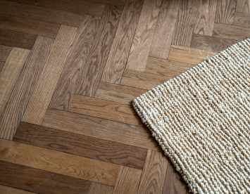 Wooden herringbone flooring in various shades of brown, bordered by a textured, light-colored woven rug.