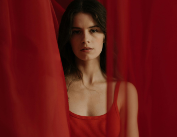 A young woman stands amidst flowing red fabric, wearing a red tank top. Her expression is serene, with soft lighting highlighting her features.