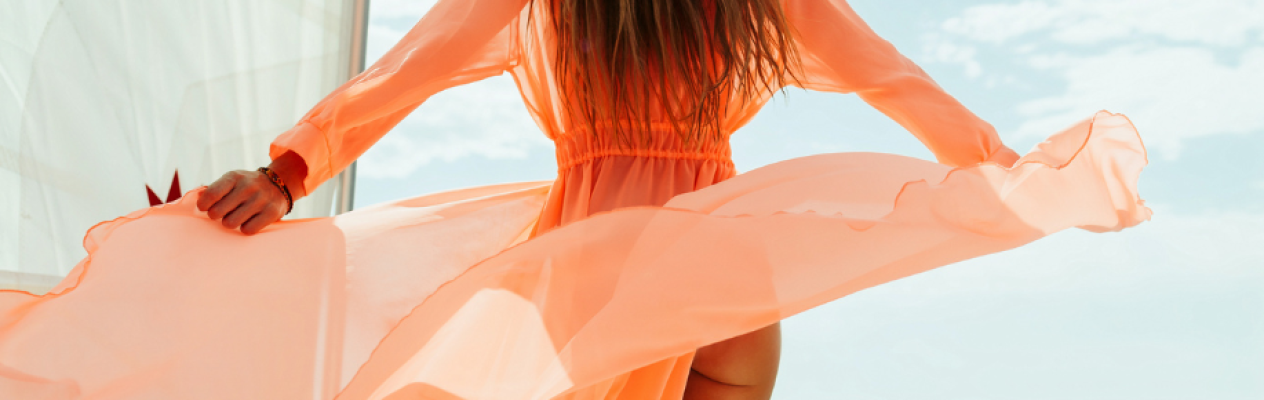 A person in a flowing, sheer orange dress stands outdoors, with the fabric billowing in the wind against a bright, sunny sky.