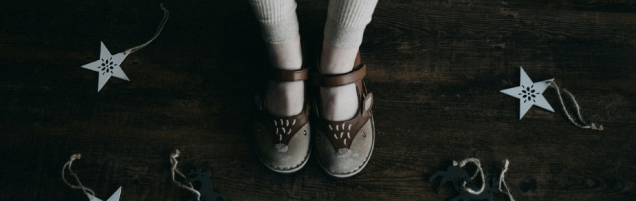 A child's feet in brown strap sandals stand on a dark wooden floor surrounded by white star-shaped decorations and other playful items.