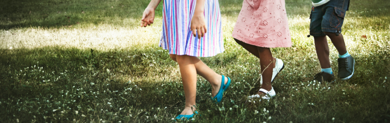 Three children walking on grass wearing colorful dresses and shorts. Wildflowers are scattered in the grass. The scene is bright and cheerful.