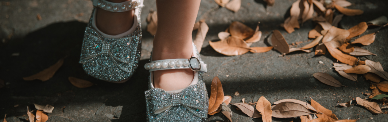 A close-up of a child's feet wearing sparkling silver shoes with a bow, standing on the ground covered with dried leaves.