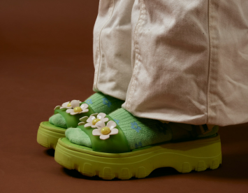 A person wearing white pants and bright green platform sandals adorned with flowers, paired with green socks featuring small blue floral patterns. The background is a warm brown color.