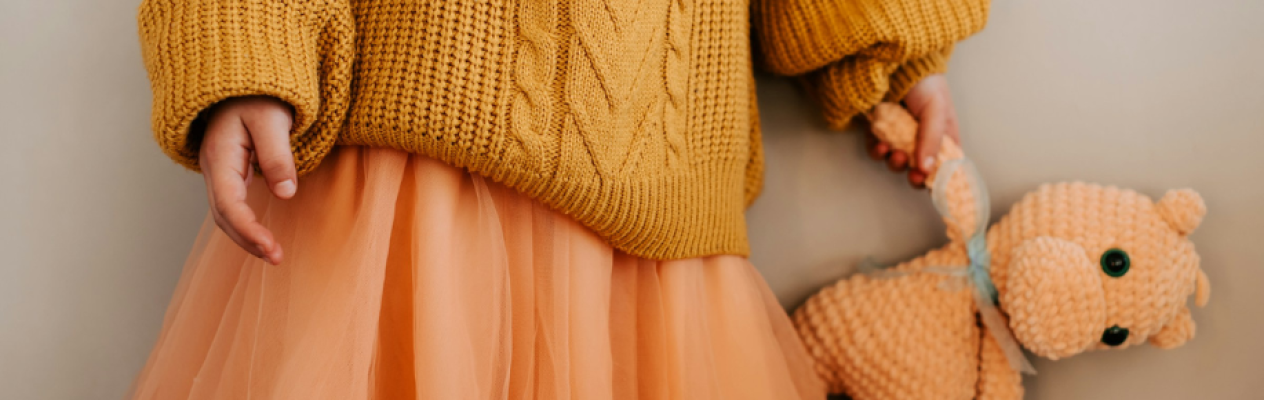 A child wearing a cozy yellow knitted sweater and a peach-colored tulle skirt holds a soft, crocheted teddy bear.