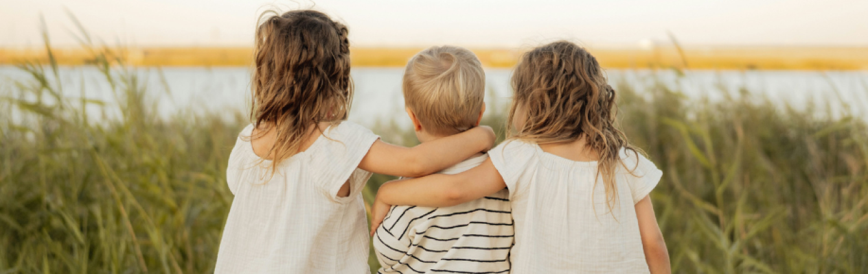Three children stand together with their backs to the camera, overlooking a body of water. Two girls, one with curly hair and another with straight hair, embrace a boy in the middle who has short, blonde hair. They are surrounded by tall grass, with a warm, golden light in the background.