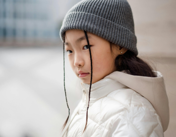 A young girl with long black hair and two braids wears a gray beanie and a light-colored puffy jacket. She looks intently at the camera with a serious expression against a blurred urban background.