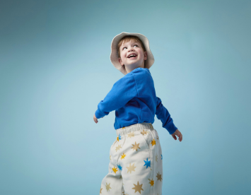 A cheerful child wearing a blue sweater and a white bucket hat looks back over their shoulder, smiling against a soft blue background. The child is dressed in light-colored pants adorned with colorful star patterns.