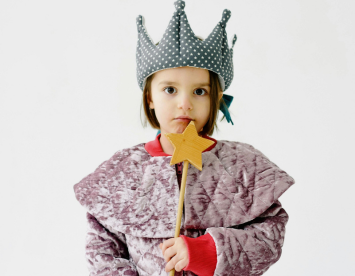 A young child wearing a polka-dotted crown and a textured, gray robe holds a wooden star wand. The child looks directly at the camera with a serious expression.